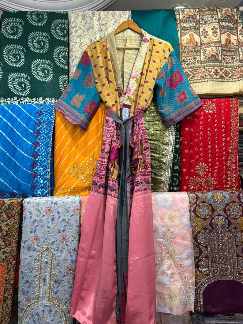 May include: A colorful, patterned kimono robe with a gray tie, hanging on a wooden hanger. The robe features a mix of yellow, pink, and blue fabrics. The background displays various patterned fabrics in different colors.