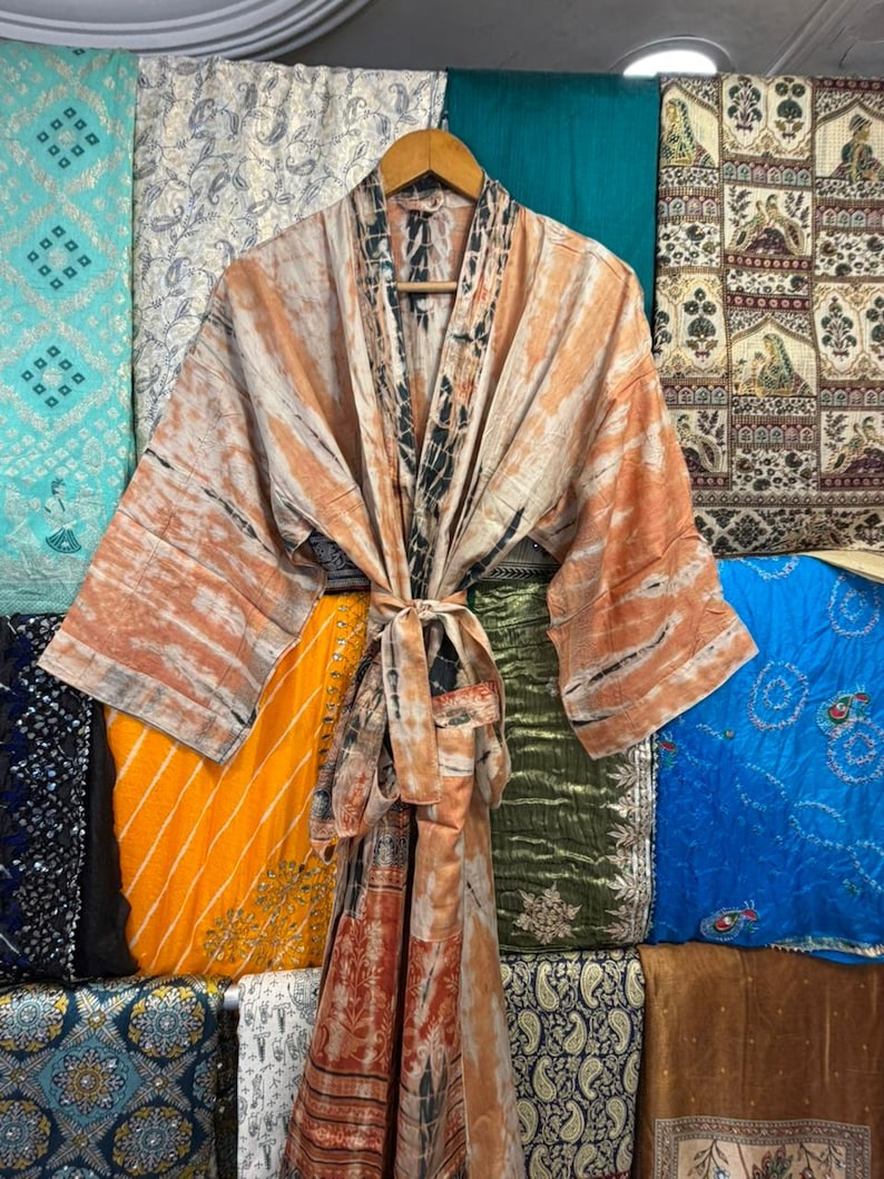 May include: A long-sleeved kimono robe featuring a tie-dye pattern in shades of peach, cream, and charcoal gray. The garment has a wide collar, an open front, and a matching fabric tie belt at the waist, displayed on a wooden hanger.