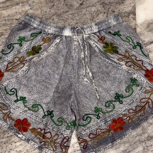 May include: Gray shorts with an elastic waistband and drawstring. The shorts are decorated with embroidered floral patterns in red, green, and gold. The edges are trimmed with white stitching.