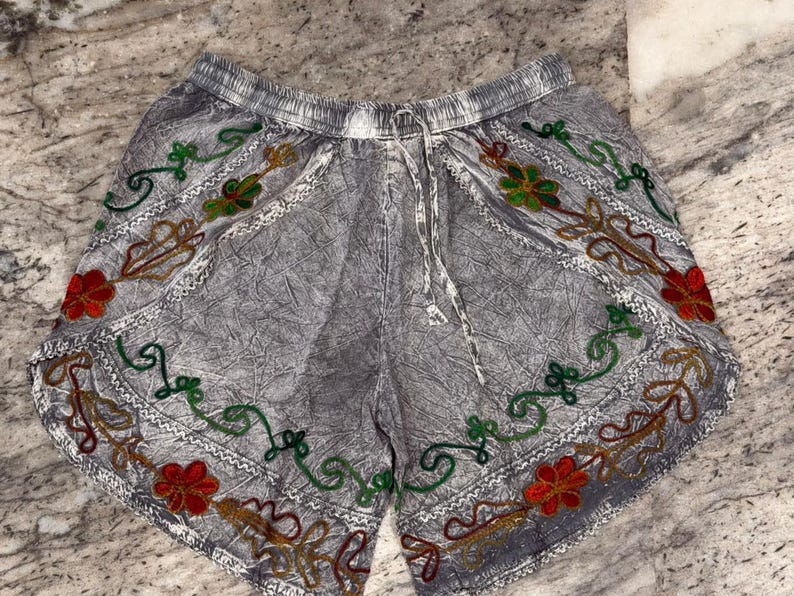 May include: Gray shorts with an elastic waistband and drawstring. The shorts are decorated with embroidered floral patterns in red, green, and gold. The edges are trimmed with white stitching.