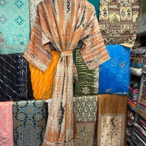 May include: A long-sleeved robe featuring a tie-dye pattern in shades of orange, white, and black. The garment includes a matching fabric belt at the waist, wide sleeves, and a wrap-front design, displayed on a wooden hanger against a textile background.