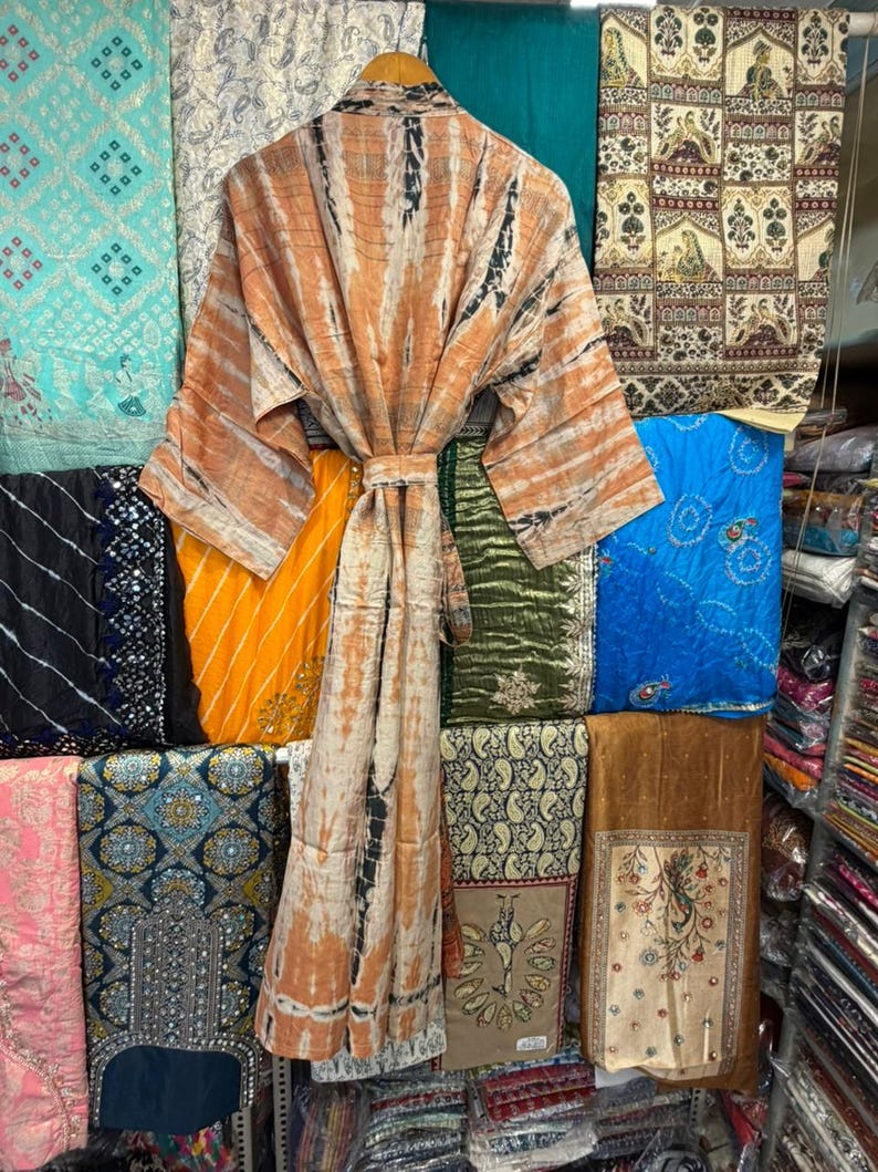 May include: A long-sleeved robe featuring a tie-dye pattern in shades of orange, white, and black. The garment includes a matching fabric belt at the waist, wide sleeves, and a wrap-front design, displayed on a wooden hanger against a textile background.