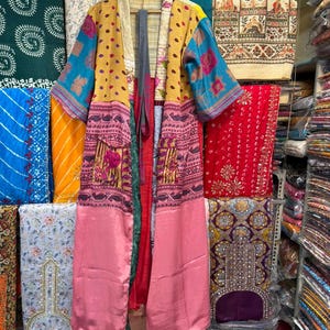 May include: A long, open-front kimono in a variety of colors and patterns. The kimono features a yellow polka-dot pattern on the upper portion, with blue and pink patterned sleeves and a pink lower section. It hangs on a wooden hanger.