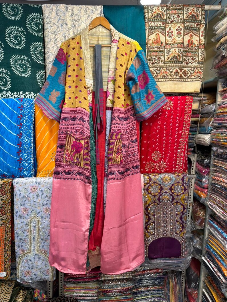 May include: A long, open-front kimono in a variety of colors and patterns. The kimono features a yellow polka-dot pattern on the upper portion, with blue and pink patterned sleeves and a pink lower section. It hangs on a wooden hanger.