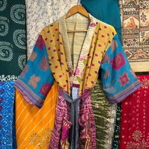 May include: A colorful, patterned kimono robe with a gray belt. The robe features a mix of yellow, blue, and pink fabrics with floral and polka dot designs. It hangs on a wooden hanger against a backdrop of various patterned textiles.