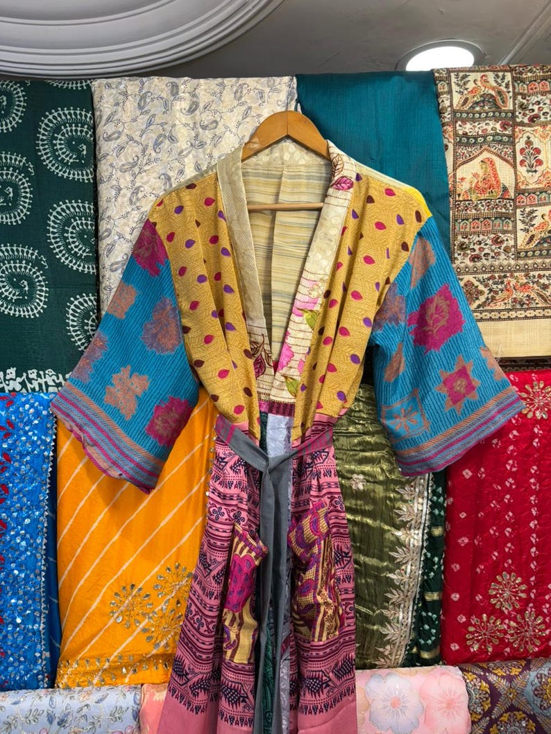 May include: A colorful, patterned kimono robe with a gray belt. The robe features a mix of yellow, blue, and pink fabrics with floral and polka dot designs. It hangs on a wooden hanger against a backdrop of various patterned textiles.