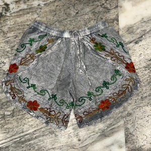 May include: Gray shorts with an elastic waistband and drawstring. The shorts are decorated with colorful embroidered floral patterns in red, green, and gold. The shorts have a relaxed fit and are made of a lightweight fabric.