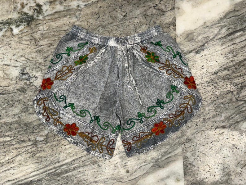 May include: Gray shorts with an elastic waistband and drawstring. The shorts are decorated with colorful embroidered floral patterns in red, green, and gold. The shorts have a relaxed fit and are made of a lightweight fabric.