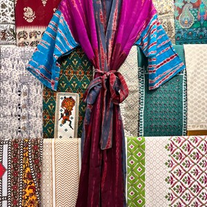 May include: A long, flowing robe with a tie belt. The robe is made of a soft, silky fabric and features a tie-dye pattern in shades of purple, blue, and gray. The robe is hanging on a hanger against a backdrop of colorful fabrics.