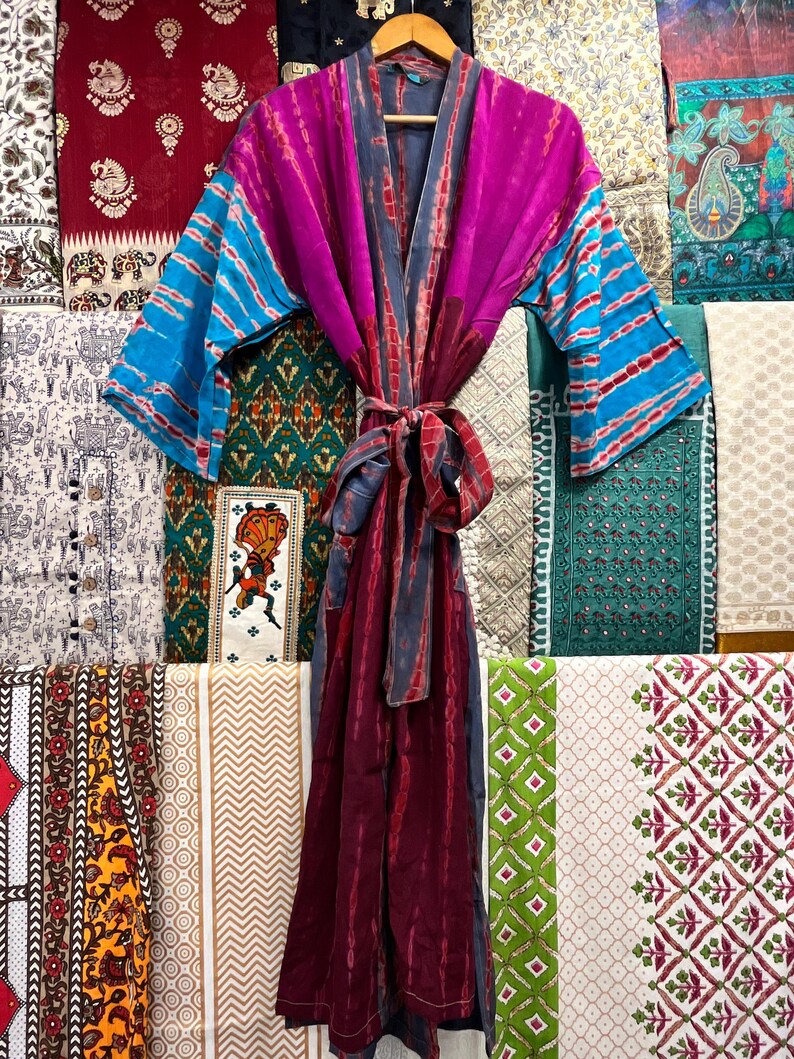 May include: A long, flowing robe with a tie belt. The robe is made of a soft, silky fabric and features a tie-dye pattern in shades of purple, blue, and gray. The robe is hanging on a hanger against a backdrop of colorful fabrics.