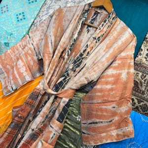 May include: A long-sleeved robe featuring a tie-dye pattern in shades of peach, cream, and black. The garment includes a matching fabric belt, wide sleeves, and a contrasting dark patterned border along the front opening and hemline.