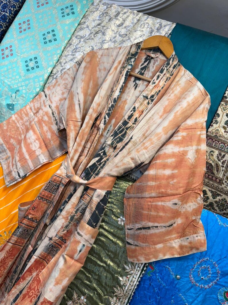 May include: A long-sleeved robe featuring a tie-dye pattern in shades of peach, cream, and black. The garment includes a matching fabric belt, wide sleeves, and a contrasting dark patterned border along the front opening and hemline.