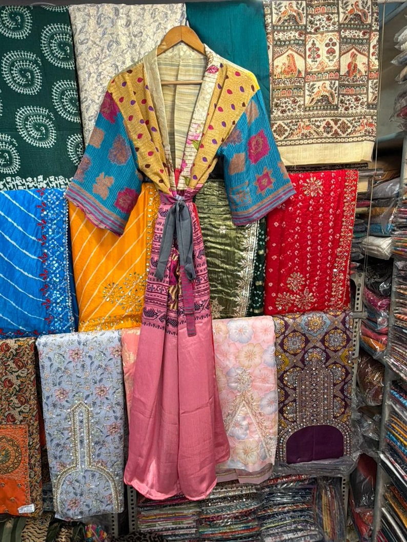 May include: A colorful, patterned kimono robe with a tie closure hangs on a wooden hanger. The robe features a mix of yellow, blue, and pink fabrics. The background displays various textiles in different colors and patterns.