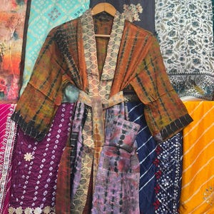 May include: A vibrant, tie-dye patterned robe with a matching belt hangs on a wooden hanger. The robe features shades of orange, brown, black, and purple. The background displays various patterned fabrics in shades of blue, purple, and yellow.