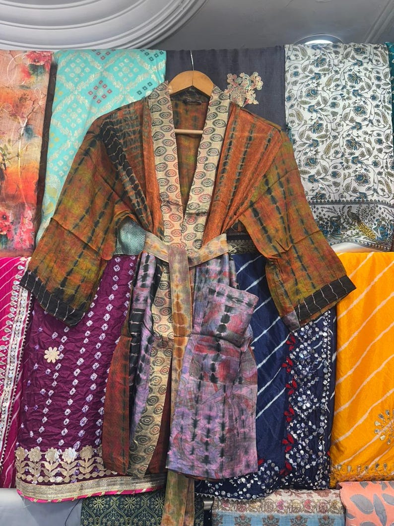 May include: A vibrant, tie-dye patterned robe with a matching belt hangs on a wooden hanger. The robe features shades of orange, brown, black, and purple. The background displays various patterned fabrics in shades of blue, purple, and yellow.
