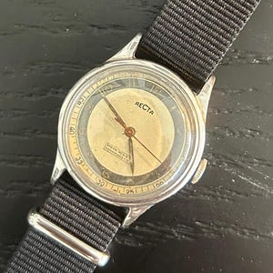 May include: Vintage RECTA wristwatch with a silver-tone case and a black nylon strap. The watch face is cream-colored with black numerals and hands. The watch has the text "RECTA" on the face. The strap is secured with a silver-tone buckle.