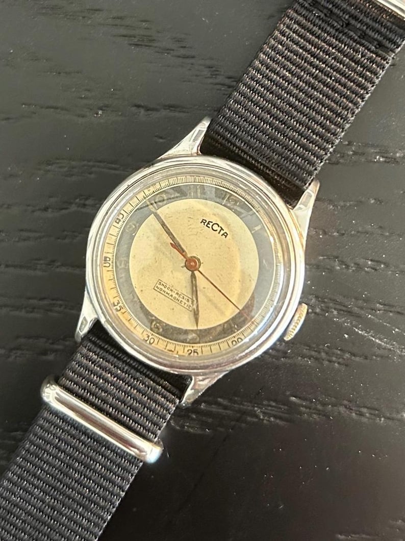 May include: Vintage RECTA wristwatch with a silver-tone case and a black nylon strap. The watch face is cream-colored with black numerals and hands. The watch has the text "RECTA" on the face. The strap is secured with a silver-tone buckle.