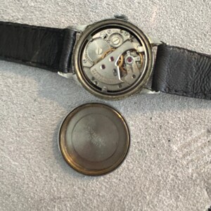 May include: An open-faced vintage wristwatch with a black leather band. The watch mechanism is visible, showcasing intricate gears, jewels, and springs. The watch's back cover is detached, revealing the inner workings. The watch is set against a neutral background.