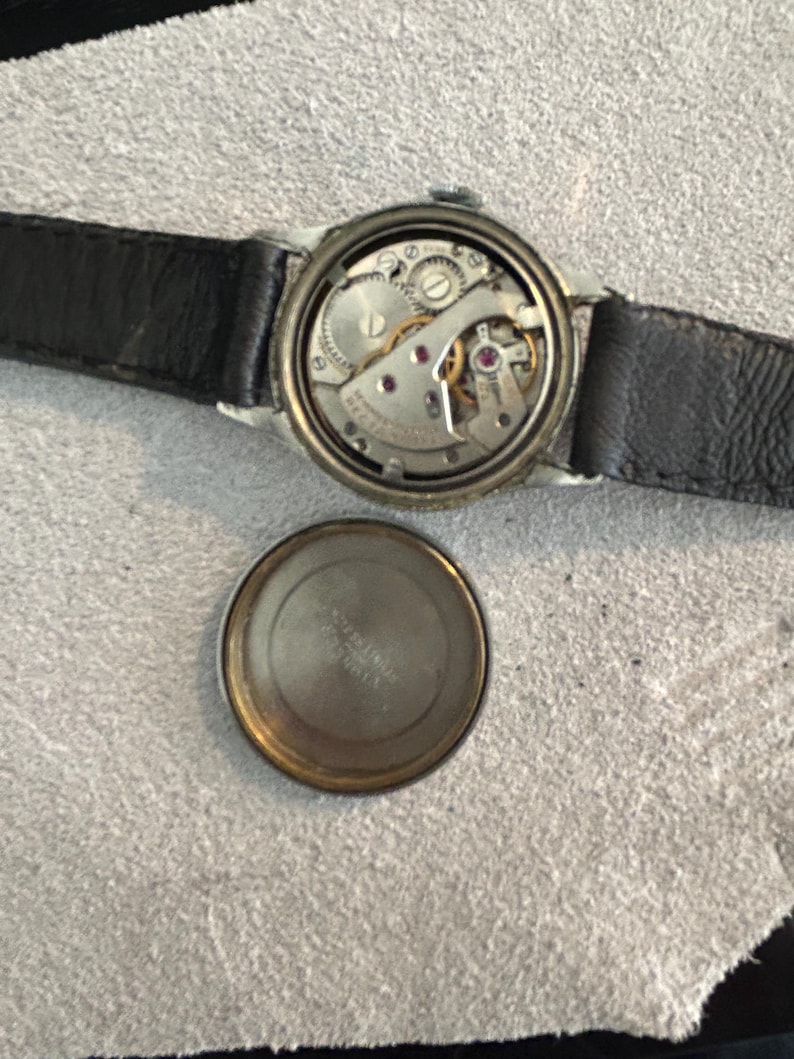 May include: An open-faced vintage wristwatch with a black leather band. The watch mechanism is visible, showcasing intricate gears, jewels, and springs. The watch's back cover is detached, revealing the inner workings. The watch is set against a neutral background.