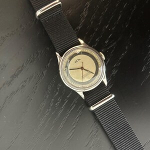May include: A vintage wristwatch with a silver-colored case and a black nylon strap. The watch face is a light beige color with black numerals and hands. The word "RECTA" is printed on the watch face.