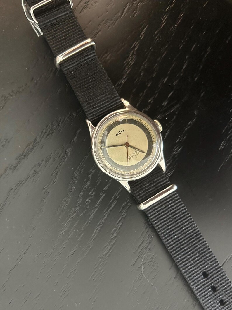 May include: A vintage wristwatch with a silver-colored case and a black nylon strap. The watch face is a light beige color with black numerals and hands. The word "RECTA" is printed on the watch face.