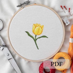 May include: A yellow tulip flower with green leaves is embroidered on white fabric in a wooden embroidery hoop.