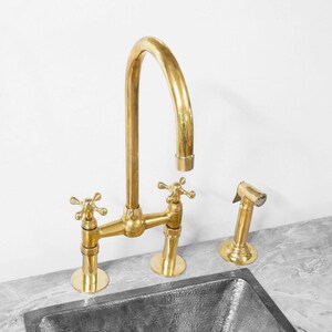 Könnte beinhalten: Eine goldfarbene Küchenarmatur mit zwei Griffen und einer seitlichen Sprühdüse. Die Armatur hat einen hohen Bogen-Auslauf und ist auf einer Arbeitsplatte montiert.