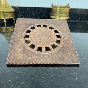 Könnte beinhalten: Ein quadratischer, dekorativer Abflussdeckel aus Messing mit einem komplizierten, gravierten Design. Der Deckel verfügt über eine kreisförmige, durchbrochene Mitte mit einem Sternmuster.