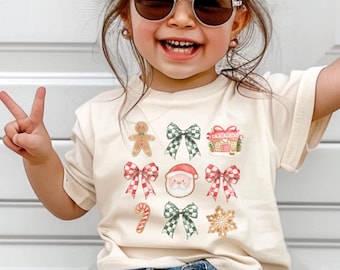 Puede incluir: Una camiseta color crema con un diseño navideño festivo. El gráfico incluye hombres de jengibre, lazos, Papá Noel y cajas de regalo. El niño lleva gafas de sol y pantalones cortos vaqueros.