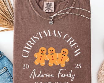 May include: A brown t-shirt with the words "CHRISTMAS CREW" in a semi-circle above three gingerbread men. Below the gingerbread men are the numbers "20" and "25", and the text "Anderson Family, New York City, NY". A silver necklace is on the shirt.