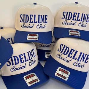 May include: A collection of baseball caps with a white crown and a blue brim. The caps feature the text "SIDELINE Social Club" in blue script. Each cap has a small tag on the brim.