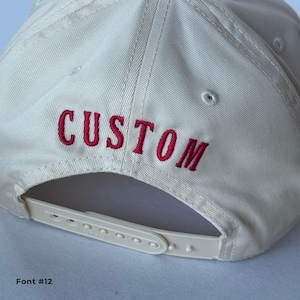 May include: A cream-colored baseball cap with the word "CUSTOM" embroidered in red thread across the back. The cap features a white adjustable strap and a curved brim. Font #12 is displayed at the bottom.