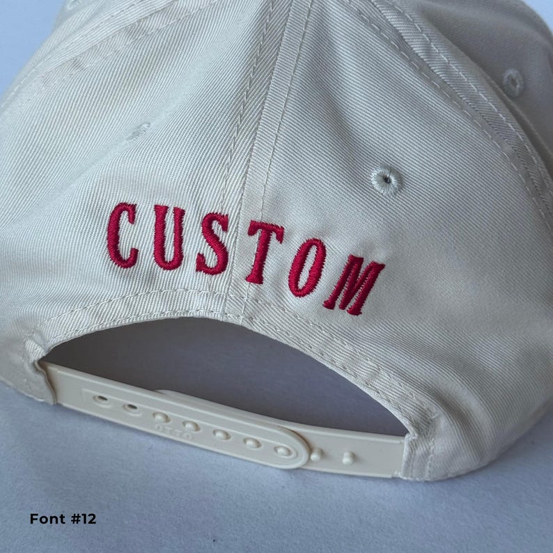 May include: A cream-colored baseball cap with the word "CUSTOM" embroidered in red thread across the back. The cap features a white adjustable strap and a curved brim. Font #12 is displayed at the bottom.