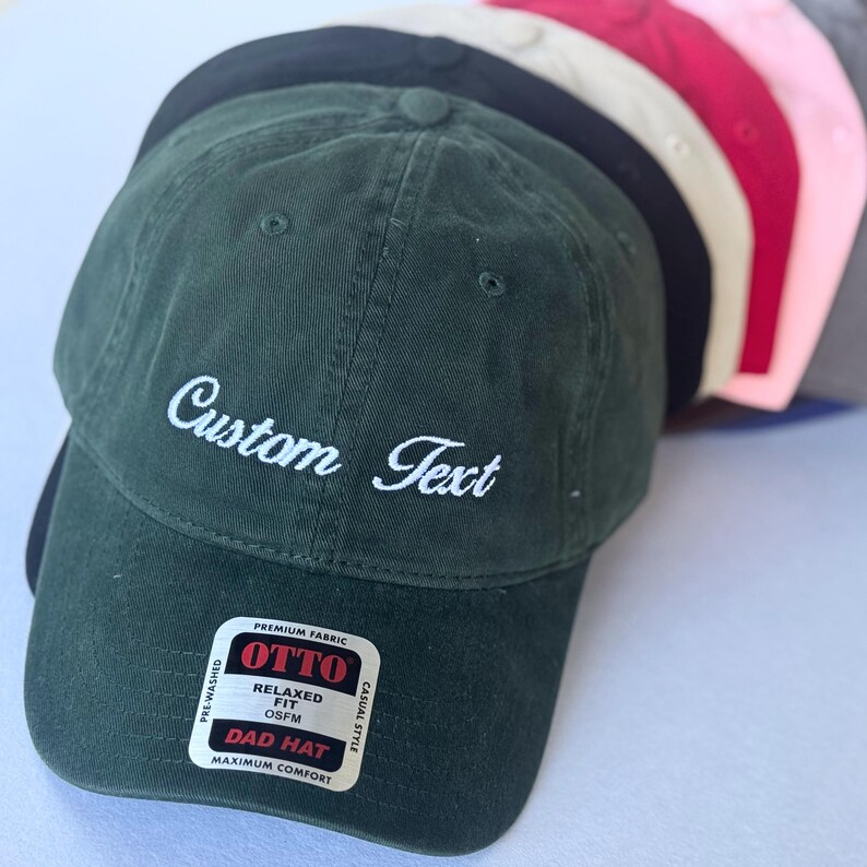 May include: A collection of pre-washed, relaxed-fit dad hats in various colors, including green, black, tan, red, pink, and gray. The green hat features the embroidered text "Custom Text" in white. The hats are made of premium fabric and are OSFM (One Size Fits Most).