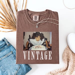 K&ouml;nnte beinhalten: Ein braunes T-Shirt mit einem Vintage-Foto eines jungen M&auml;dchens, das die Kerzen auf einem Geburtstagskuchen ausbl&auml;st. Der Text "Vintage" ist in Wei&szlig; unter dem Foto aufgedruckt.