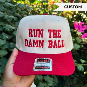 May include: A white and red baseball cap with the text "RUN THE DAMN BALL" embroidered in red. The cap has a classic fit and a mid profile. The word "CUSTOM" is in a white banner.