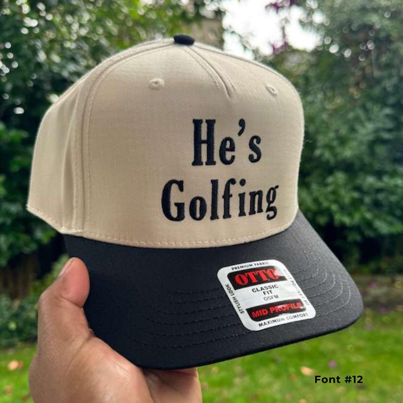 May include: A beige and black baseball cap with the text "He's Golfing" in black. The cap has a classic fit and a mid-profile design. The cap is made of premium fabric.