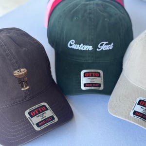 May include: A collection of baseball caps in various colors. One cap is dark brown with an embroidered cocktail glass, another is dark green with "Custom Text" in white, and a third is beige. Each cap has a "DAD HAT" label.