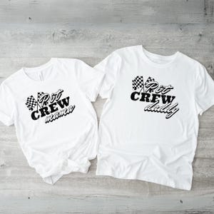 May include: Two white t-shirts with black checkered flags and text. One shirt says "Pit Crew mama" and the other says "Pit Crew daddy".