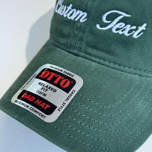 May include: A dark green Dad Hat with white embroidered text that reads "Custom Text". The hat has a curved brim and a tag that says "OTTO" and "Relaxed Fit". The hat is pre-washed and made of premium fabric.