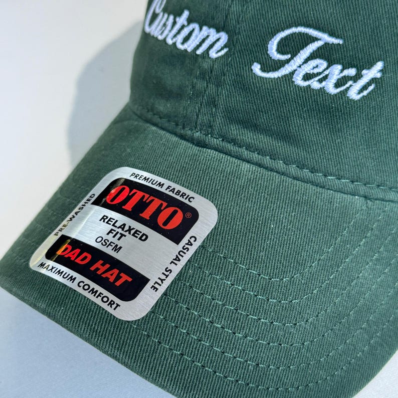 May include: A dark green Dad Hat with white embroidered text that reads "Custom Text". The hat has a curved brim and a tag that says "OTTO" and "Relaxed Fit". The hat is pre-washed and made of premium fabric.