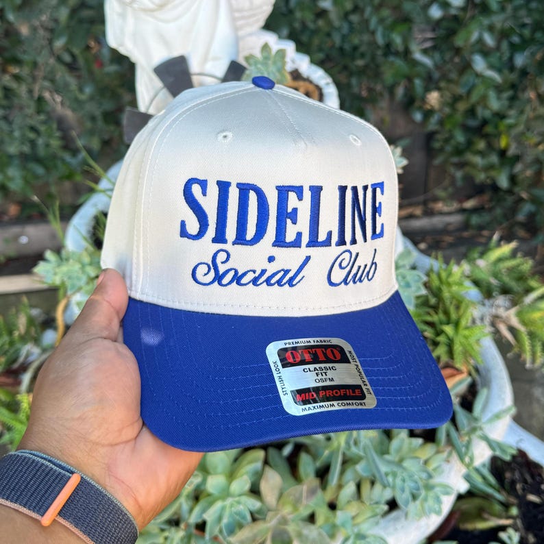 May include: A white and blue baseball cap with the text "SIDELINE Social Club" embroidered in blue. The cap has a blue brim and a blue button on top. The cap is made of a premium fabric.
