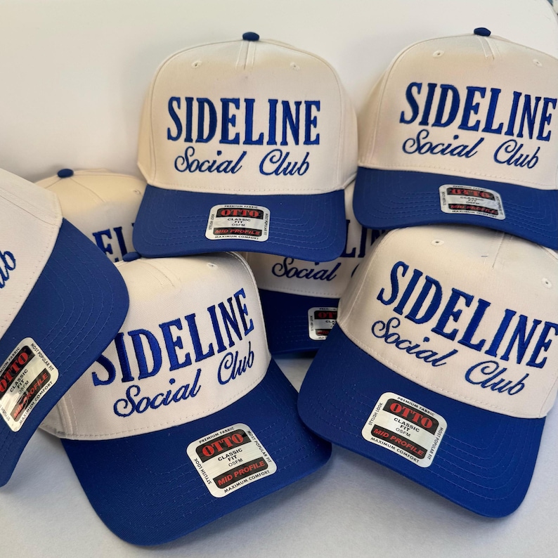 May include: A collection of baseball caps with a white crown and a blue brim. The caps feature the text "SIDELINE Social Club" in blue script. Each cap has a small tag on the brim.