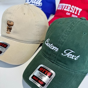 May include: A collection of baseball caps in various colors. One tan cap features an embroidered cocktail design. A green cap displays "Custom Text" in white script. Other caps are red and blue, with "University" and "Dad" printed on them.