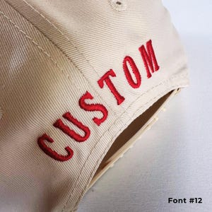 May include: A beige baseball cap with the word "CUSTOM" embroidered in red on the side. The text is in a bold, stylized font. The image also shows the text "Font #12" in the bottom right corner.