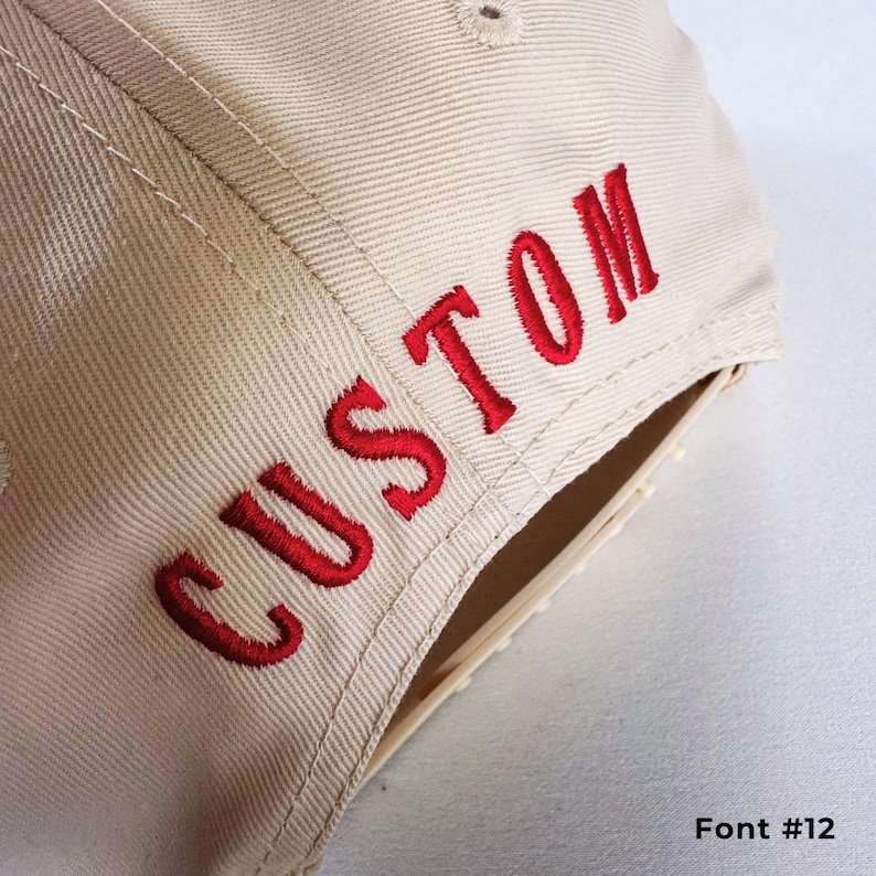May include: A beige baseball cap with the word "CUSTOM" embroidered in red on the side. The text is in a bold, stylized font. The image also shows the text "Font #12" in the bottom right corner.