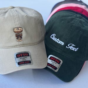 May include: A collection of baseball caps in various colors. One tan cap features an embroidered cocktail design. A dark green cap displays "Custom Text" in white script. Each cap has a "Dad Hat" label.