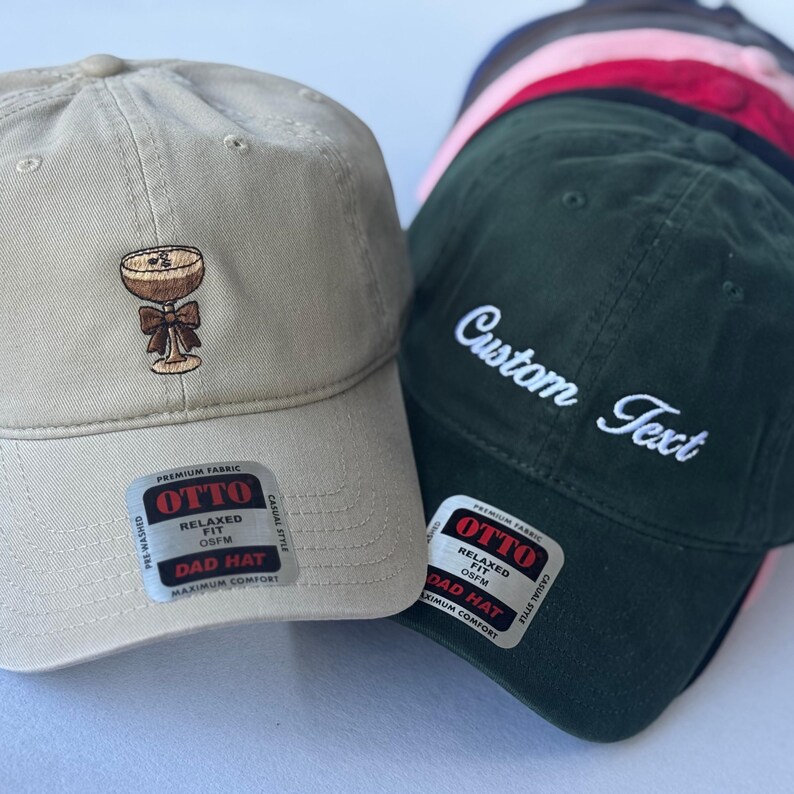 May include: A collection of baseball caps in various colors. One tan cap features an embroidered cocktail design. A dark green cap displays "Custom Text" in white script. Each cap has a "Dad Hat" label.