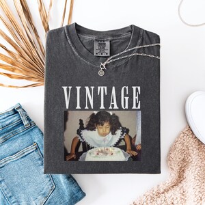 K&ouml;nnte beinhalten: Ein Vintage-T-Shirt in Grau mit dem in Wei&szlig; gedruckten Wort "Vintage". Das Shirt zeigt ein Foto eines jungen M&auml;dchens, das die Kerzen auf einem Geburtstagskuchen ausbl&auml;st.