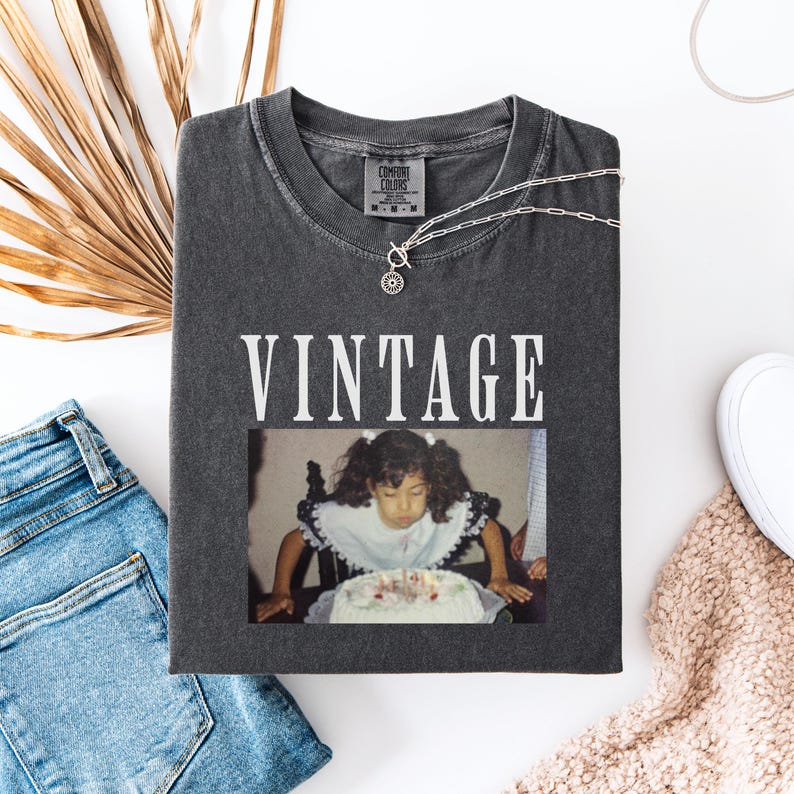 K&ouml;nnte beinhalten: Ein Vintage-T-Shirt in Grau mit dem in Wei&szlig; gedruckten Wort "Vintage". Das Shirt zeigt ein Foto eines jungen M&auml;dchens, das die Kerzen auf einem Geburtstagskuchen ausbl&auml;st.